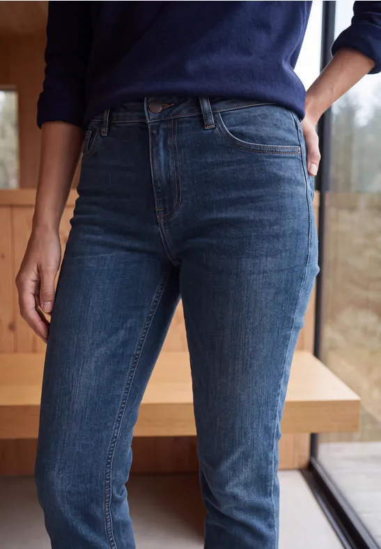 Une personne porte un jean taille haute bleu foncé, debout près d’une fenêtre devant un mur en bois.