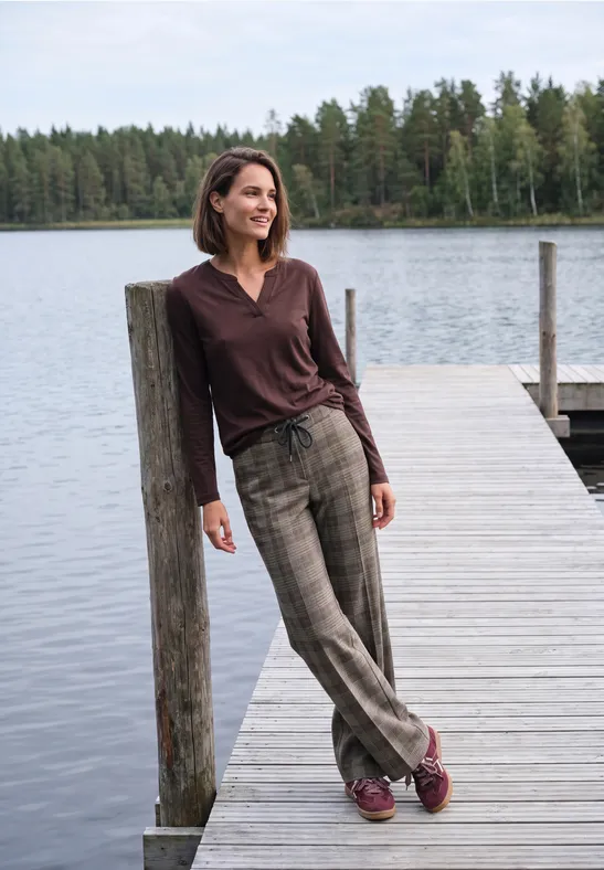Une femme appuyée à un poteau sur un ponton, devant un lac et une forêt.