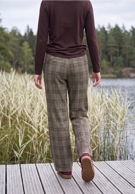 Personne en pantalon à carreaux et manches longues sur un ponton au bord d’un lac, entourée de roseaux et forêt.