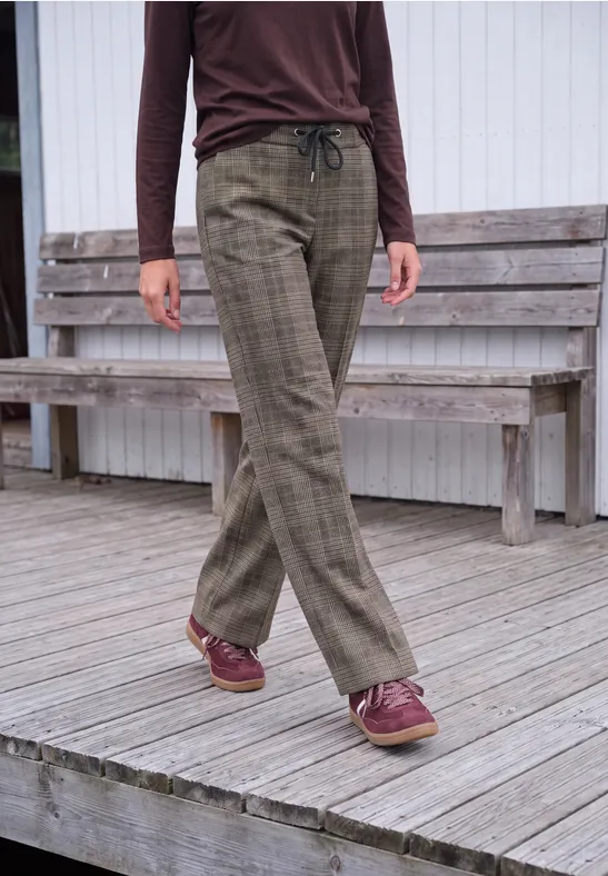 Femme en haut marron, pantalon olive à carreaux et chaussures bordeaux sur terrasse en bois.