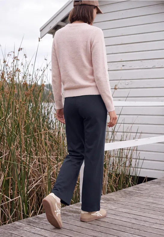 Une femme en pull beige et pantalon sombre marche sur une jetée près des roseaux.