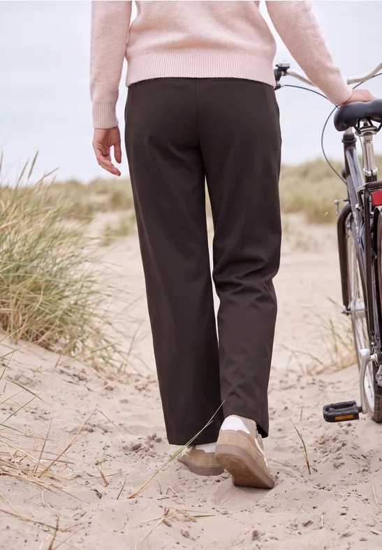Une personne pousse un vélo dans les dunes, vêtue d'un pantalon noir et d'un pull rose.