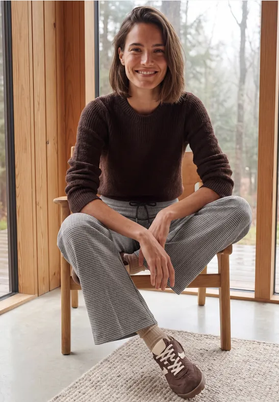 Femme souriante assise sur une chaise face à une grande fenêtre dans une pièce claire et moderne.
