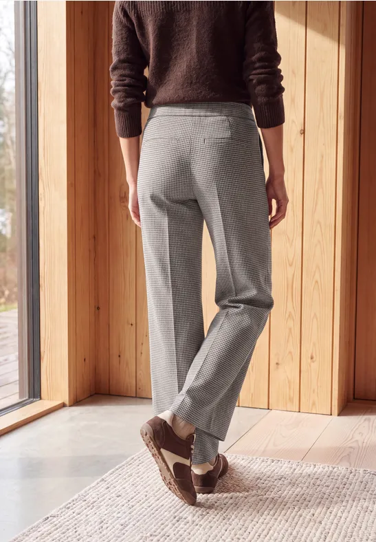 Personne en pantalon gris et chaussons, vue arrière, debout devant une grande fenêtre dans un intérieur en bois clair.