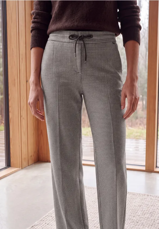 Une personne porte un pantalon gris droit à cordon de taille et un pull marron devant de grandes fenêtres.