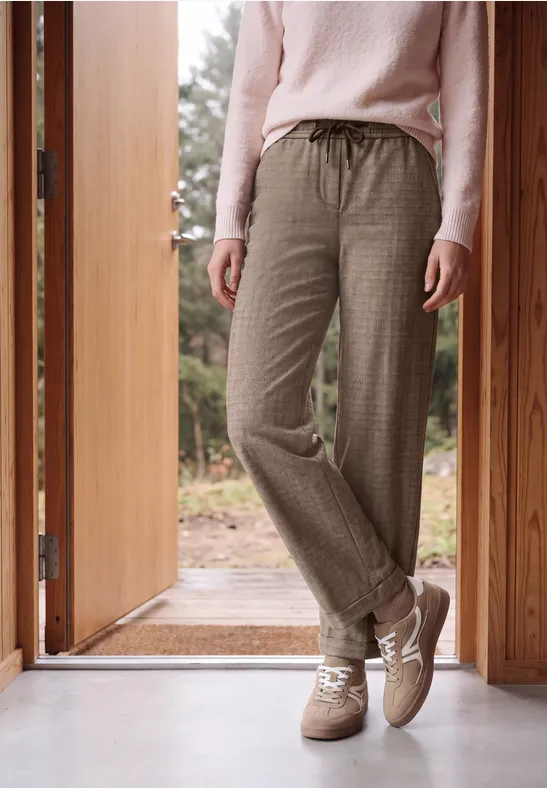 Personne debout dans l'encadrement d'une porte, portant un pull clair, un pantalon marron à carreaux et des baskets.