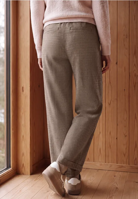 Une personne vue de dos porte un pantalon en laine beige, un pull rose et des baskets dans un intérieur en bois.