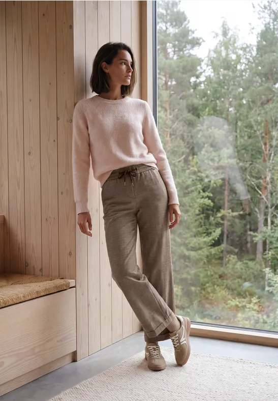 Une femme en pull rose et pantalon beige se tient pieds nus près d’une fenêtre dans une pièce lumineuse face à la nature.