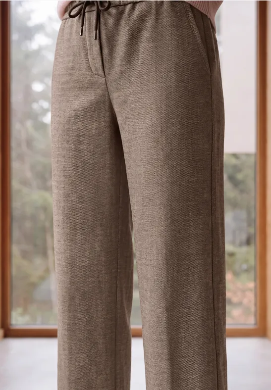 Une personne porte un pantalon ample beige-brun à cordon devant une grande fenêtre.