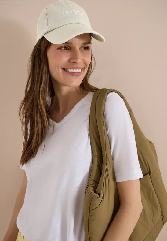 Personne en T-shirt blanc et casquette claire porte un sac matelassé vert olive sur l’épaule.