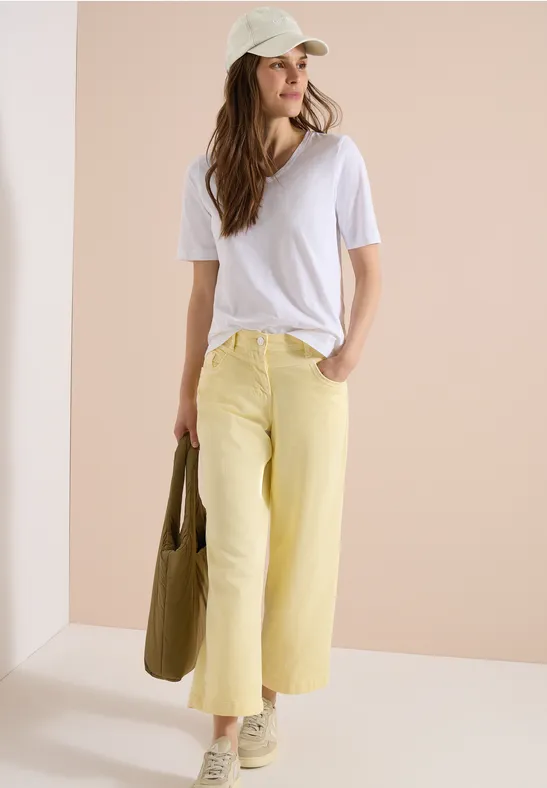 Femme en t-shirt blanc, pantalon large jaune pastel, casquette et sac vert sur fond clair.