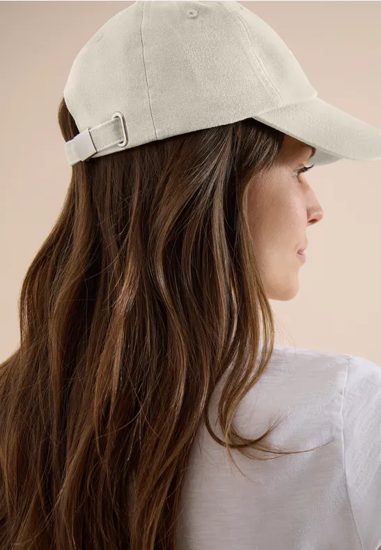 Femme de dos aux cheveux bruns longs, portant un t-shirt blanc et une casquette beige sur fond beige.