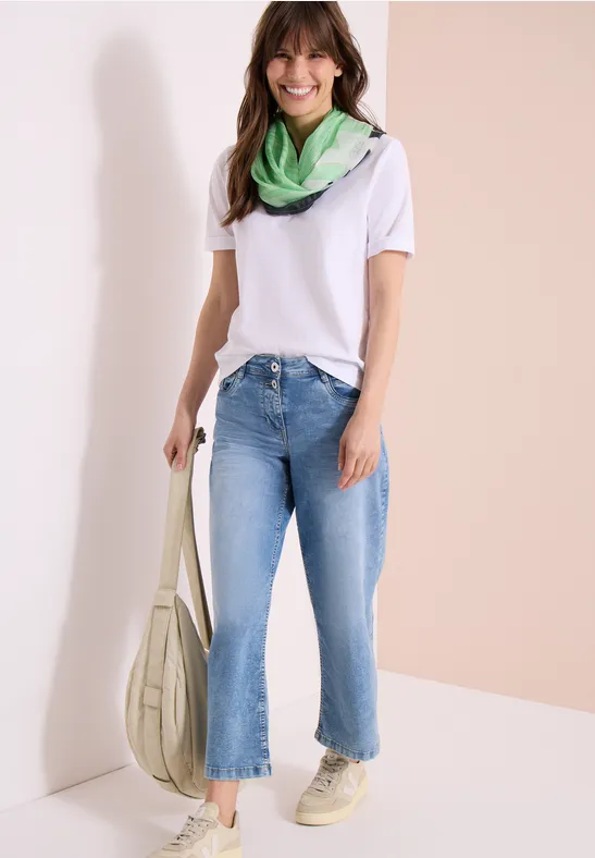 Frau in weißem T-Shirt, grünem Halstuch, blauen Jeans und Sneakern mit cremefarbener Tasche.