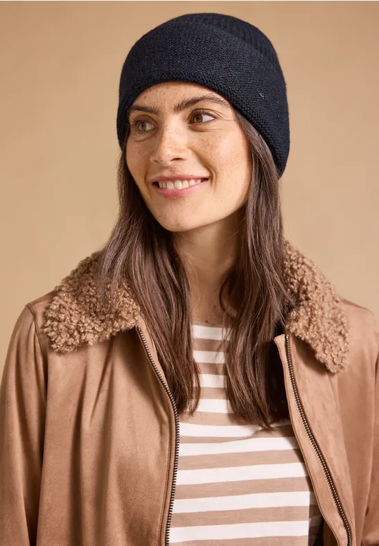 Frau mit schwarzer Strickmütze, brauner Jacke mit Teddy-Kragen und gestreiftem Shirt vor beigem Hintergrund.