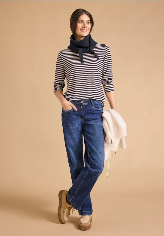 Eine Frau trägt eine gestreifte Bluse, Jeans, Halstuch, Halbschuhe und hält eine Jacke lässig in der Hand.
