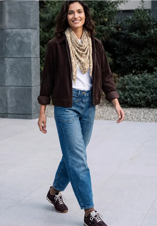 Eine fröhliche junge Frau in Jeans, weißem Top und brauner Jacke mit Schal spaziert auf einem modernen Gehweg.