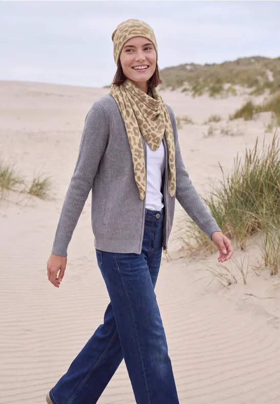 Eine lächelnde Frau spaziert am Dünenstrand, trägt Jeans, graue Strickjacke und passende Mütze mit Schal.