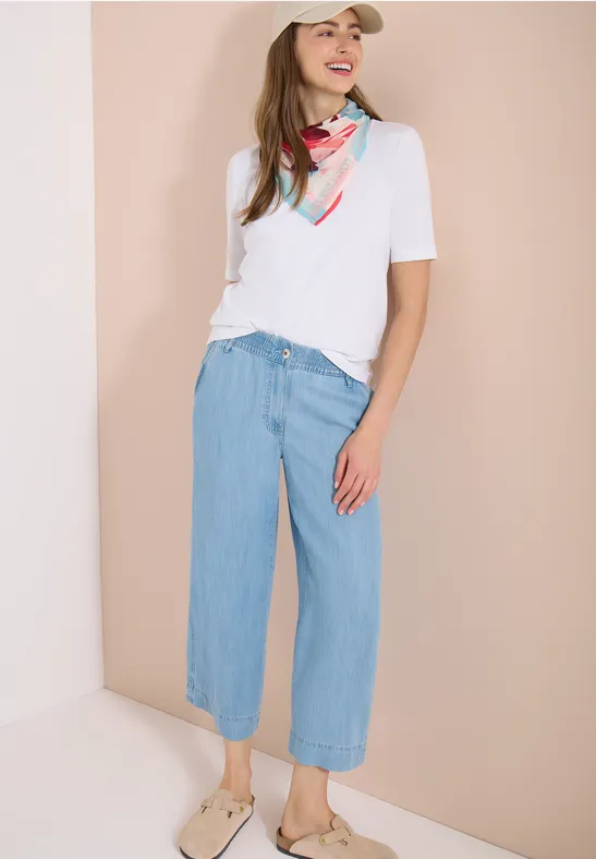 Frau mit weißem T-Shirt, buntem Halstuch, weiten hellblauen Jeans-Culottes und Sandalen vor hellem Hintergrund.