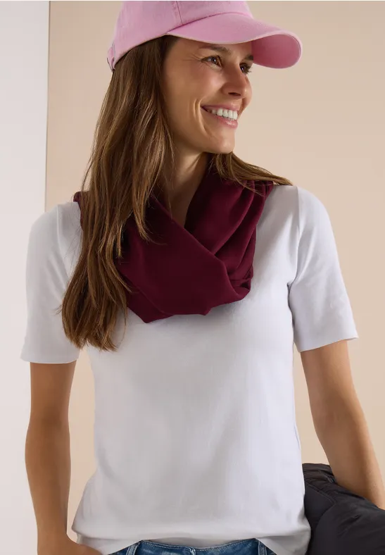 Eine lächelnde Frau in weißem T-Shirt, rosa Cap und weinrotem Schal über den Schultern, hält eine Jacke.
