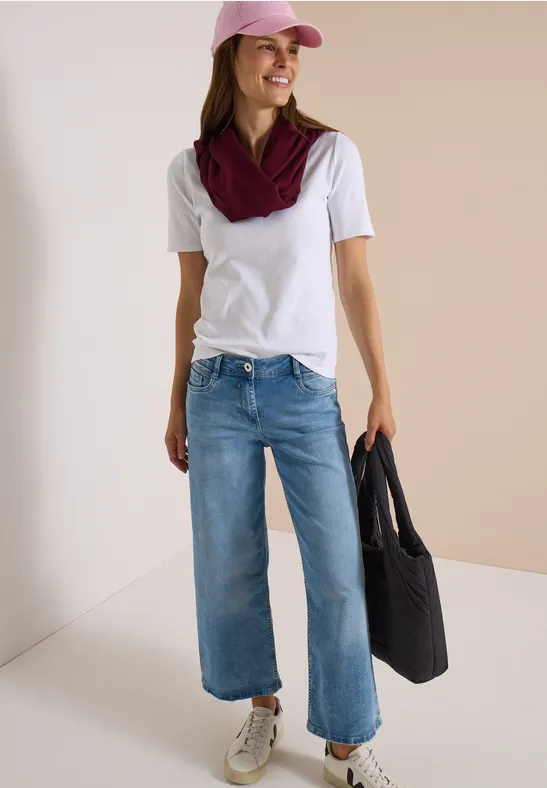 Frau in weißem Shirt, rosa Cap, burgunderfarbenem Schal, weit geschnittenen Jeans, weißen Sneakers und schwarzer Tasche.