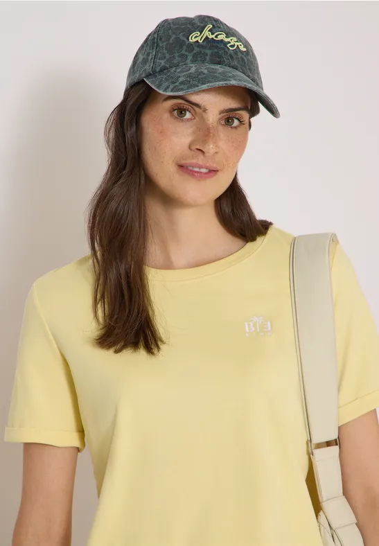 Femme souriante portant une casquette en jean, un t-shirt jaune et un sac à l'épaule.