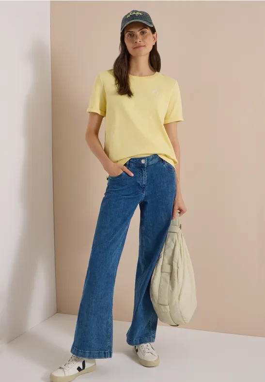 Femme en t-shirt jaune, jean large, casquette verte et sac blanc devant un mur clair.