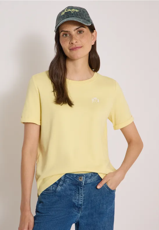 Une femme souriante portant un t-shirt jaune, un jean bleu et une casquette devant un fond neutre.