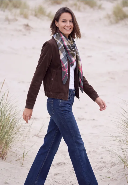 Vrouw wandelt lachend blootsvoets in jeans, bruine jas en kleurrijke sjaal over duinen
