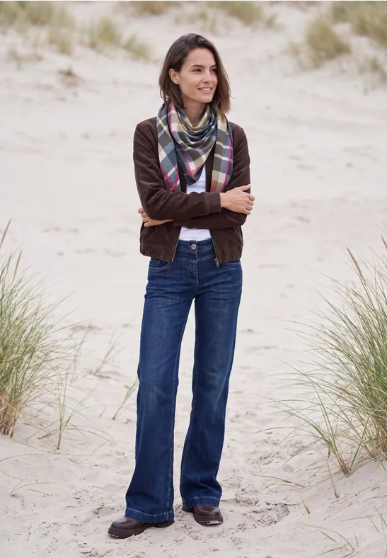 Een lachende vrouw in jeans, jasje en sjaal met gekruiste armen op zandduinen.
