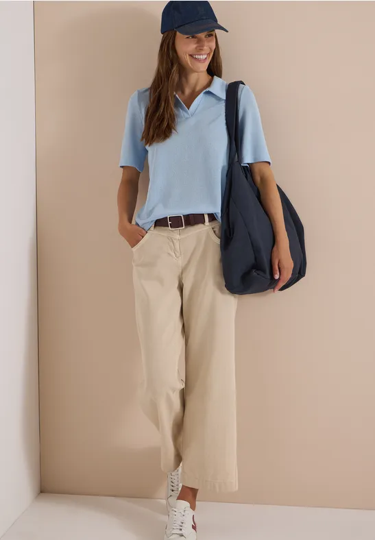 Femme en polo bleu clair, pantalon beige, baskets blanches, casquette bleue et grand sac à bandoulière.