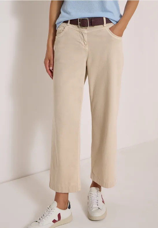 Femme en pantalon beige ample à jambe droite et baskets blanches devant un mur clair.