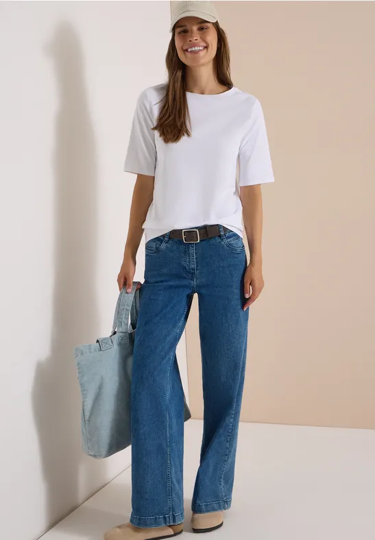 Une femme souriante porte un t-shirt blanc, un jean large bleu avec ceinture et tient un sac en jean clair.