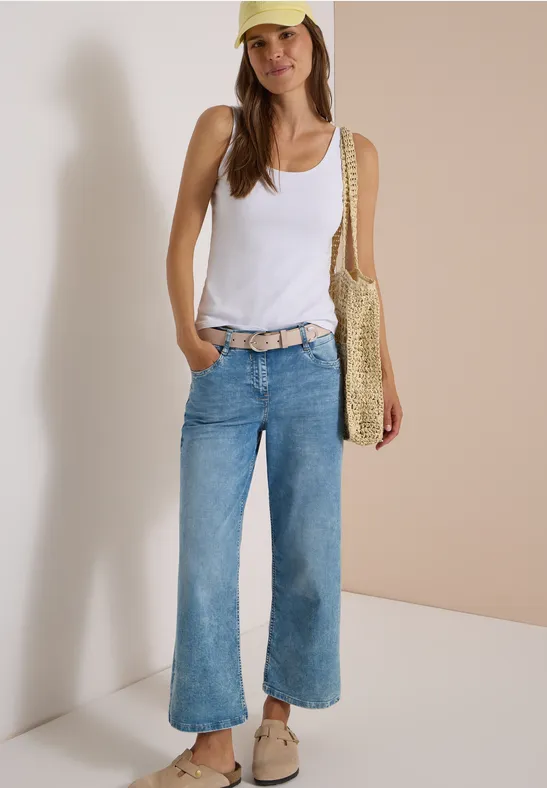 Une femme porte un débardeur blanc, jean large, ceinture beige, chaussures beiges, sac en paille et casquette jaune.