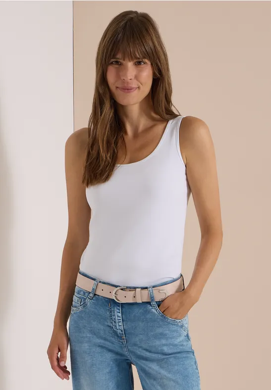 Jeune femme souriante en débardeur blanc et jean avec ceinture rose-gris, fond neutre.