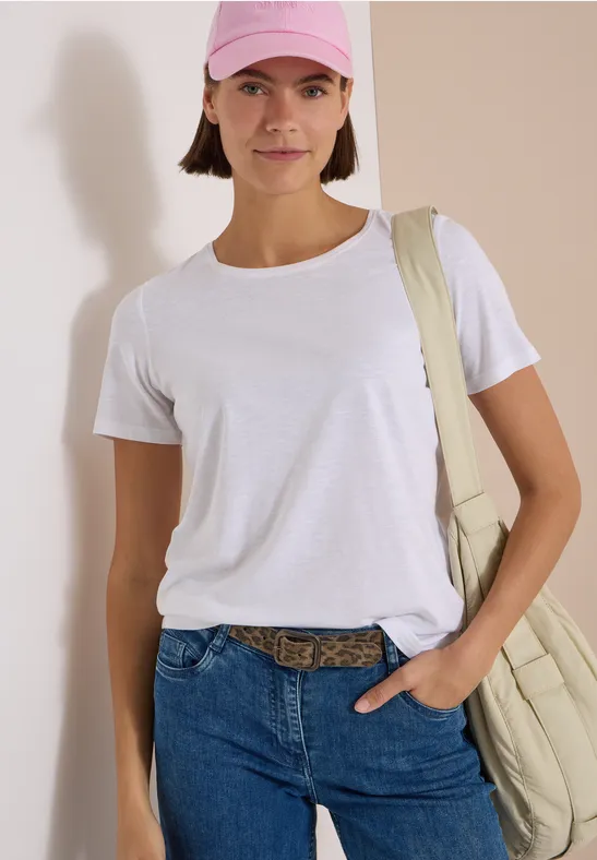 Femme en t-shirt blanc, jean et casquette rose, sac épaule, debout devant un mur clair.