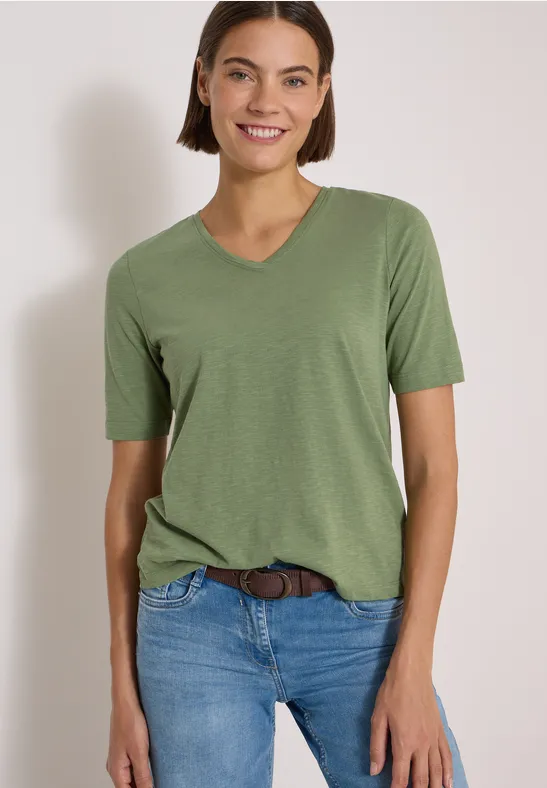 Femme souriante en T-shirt vert à encolure en V et jean, fond gris clair.