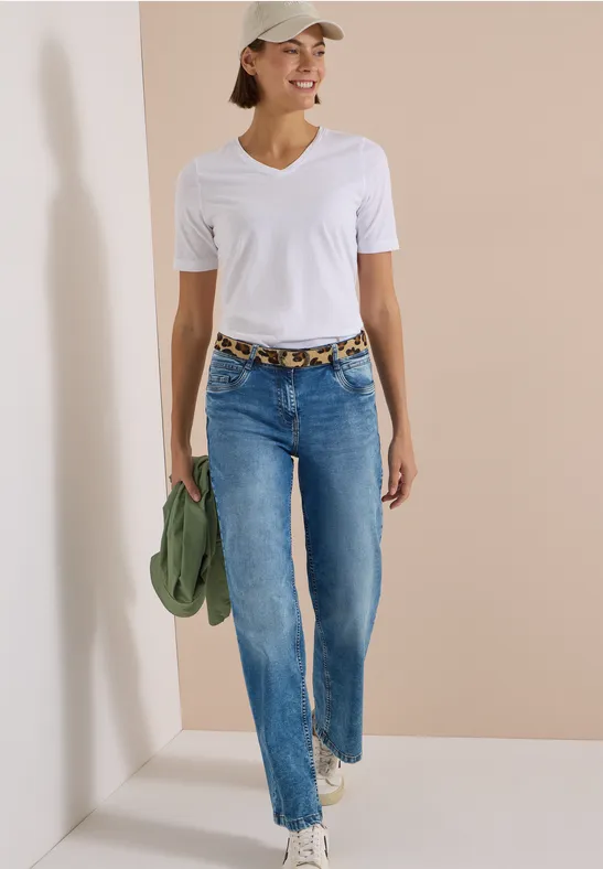 Femme souriante en t-shirt blanc, jean et casquette, ceinturée léopard, tenant une veste verte et baskets.