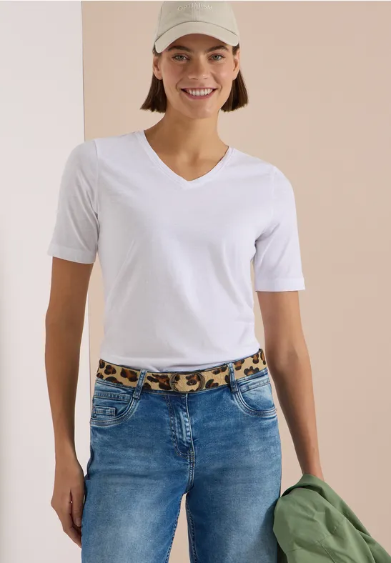 Femme souriante en T-shirt blanc à col en V et jean avec ceinture léopard, casquette beige et veste verte.