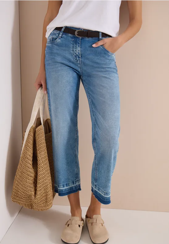 La personne porte un jean large bleu, ceinture, haut blanc, sac en osier et mules claires.