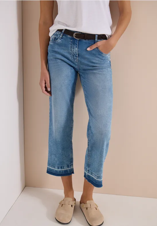Personne porte un jean droit bleu clair et des mocassins beiges sur fond neutre.