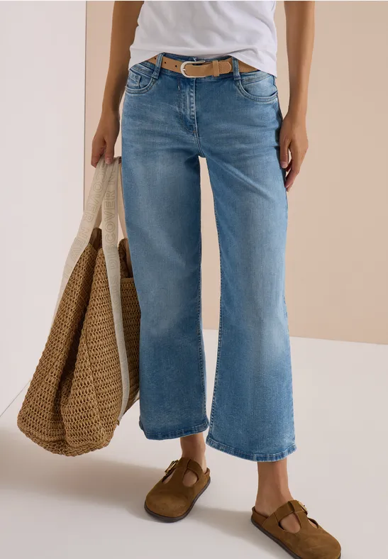 Personne porte un t-shirt blanc, un jean large bleu clair, des chaussures marron et tient un sac en paille.