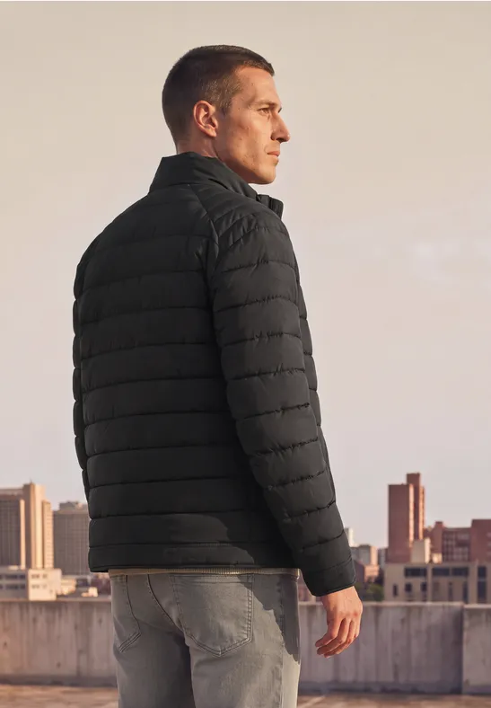 Ein Mann in schwarzer Steppjacke steht auf einem Dach und blickt in die Ferne vor urbaner Skyline.