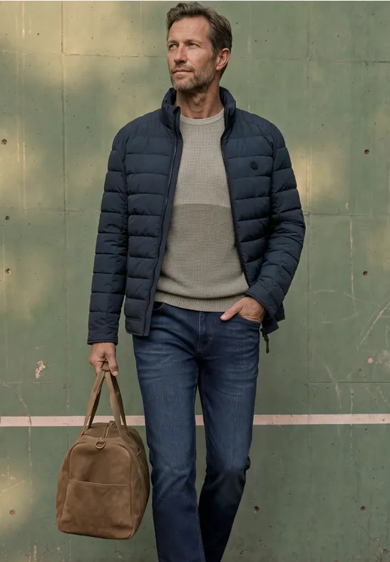 Mann in gesteppter Jacke, Jeans und Pullover trägt eine braune Reisetasche vor grüner Wand.