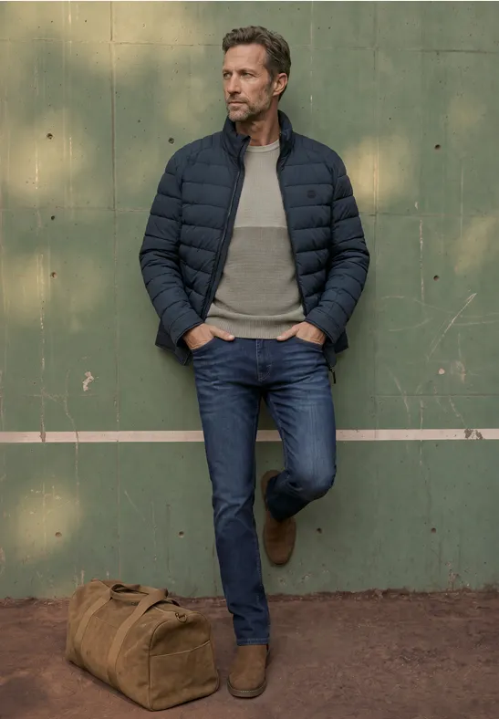 Ein lässig gekleideter Mann lehnt in Jeans und Daunenjacke vor grüner Wand, Reisetasche neben ihm.