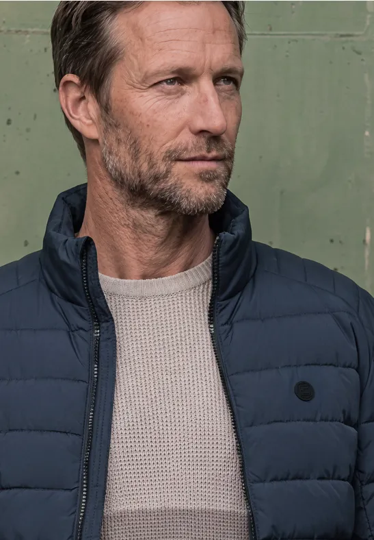 Ein ernst blickender Mann um die 40 mit grauem Bart trägt eine dunkelblaue Steppjacke vor grünem Hintergrund.