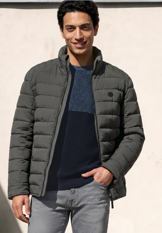 Ein junger Mann steht lässig in einer grauen Steppjacke über dunkelblauem Pullover und Jeans.