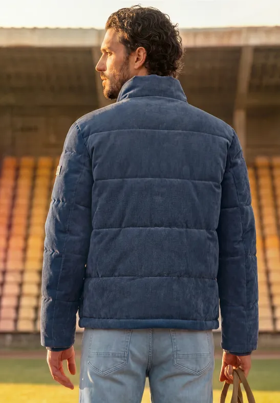 Ein Mann in einer blauen Steppjacke steht mit dem Rücken zur Kamera und hält eine Tasche im Stadion.