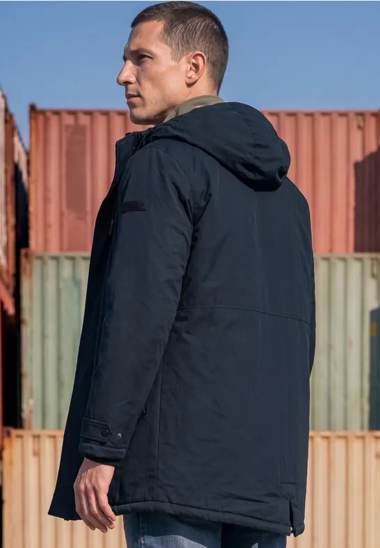 Un homme en parka sombre à capuche se tient de profil devant des conteneurs colorés.