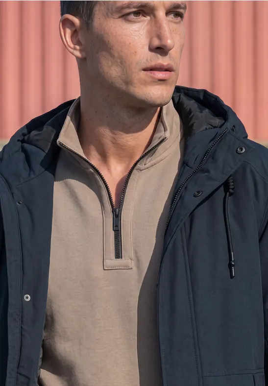 Un homme en shirt beige à demi-zip et veste bleu foncé devant un mur rose.