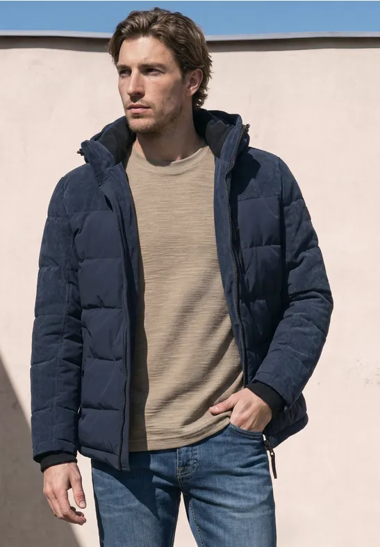 Ein Mann trägt eine dunkelblaue Steppjacke, einen beigen Pullover und Jeans vor hellem Hintergrund.